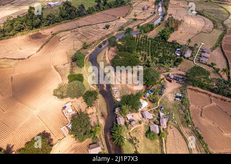 Vista aérea de tranquilo pueblo local con río que fluye entre el valle y la plantación de arroz en temporada seca en el campo de Muang Kong, Chia