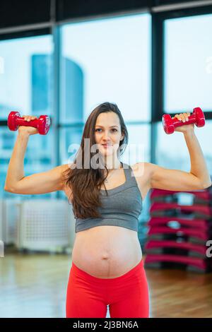 Retrato de una mujer embarazada de pelo oscuro que hace ejercicios aeróbicos en un estudio deportivo cerca de la ventana con mancuernas rojas. Vista lateral del día soleado. Bolas de ajuste rosas A Foto de stock