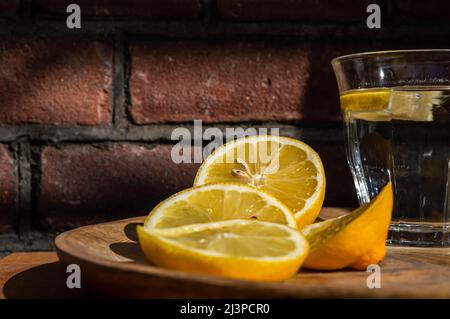 Imagen de rodajas de limón y un vaso de agua en un plato de madera al sol Foto de stock