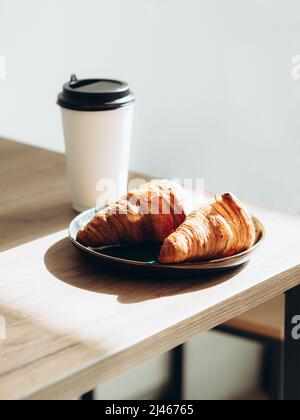 Taza de café para llevar en una taza de papel blanco y croissants a la luz del sol sobre una mesa de madera. Café por la mañana en el café para texto. Foto de stock