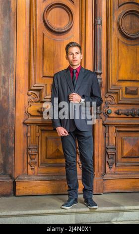 Vestirse en una camiseta interior roja, un blazer negro, jeans negros y una  corbata negra, un estudiante universitario se inclina contra una columna  fuera de una oficina en el campu Fotografía de