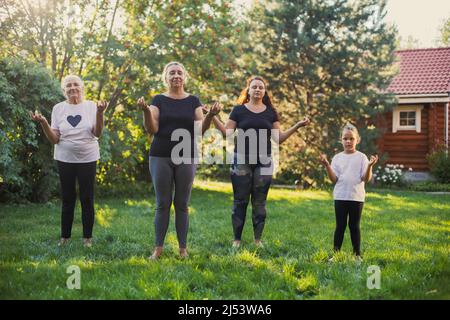 Mujeres brillantes Cuatro generaciones de familia haciendo yoga meditando con sus manos levantadas y los dedos conectados de pie en la pradera llena de vegetación Foto de stock