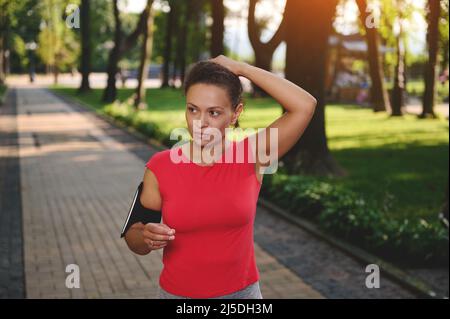 Determinada deportista de mediana edad en una camiseta roja con soporte para smartphone atado con cola de caballo mientras se encuentra en el camino por la mañana al correr a lo largo del parque. Cardio w Foto de stock