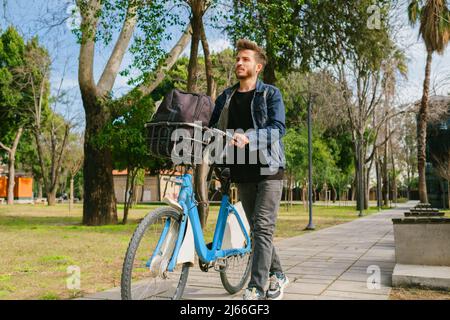 Foto de perfil completo de un hombre con ropa vaquera caminando y empujando una bicicleta sobre el verde fondo del parque de la ciudad. Hombre llevándole la mochila en bicicleta. Foto de stock
