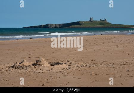 Un lejano Castillo de Dunstanburgh visto desde la playa de Embleton en un día de verano con un castillo de arena de un niño con foso actuando como un punto de vista en primer plano Foto de stock
