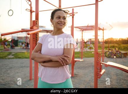 Retrato seguro de una agradable mujer multiétnica de mediana edad, atleta, deportista en activo posando con brazos cruzados contra el crossbar en el sp Foto de stock