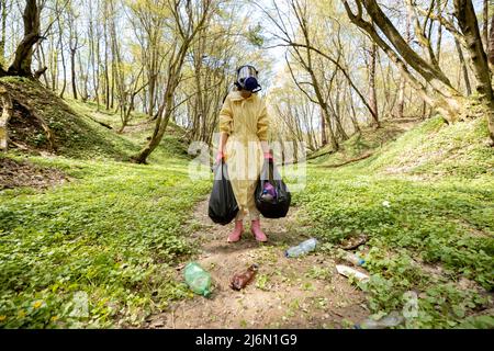 Mujer con máscara de gas y ropa protectora recolectando basura plástica dispersa en el bosque Foto de stock
