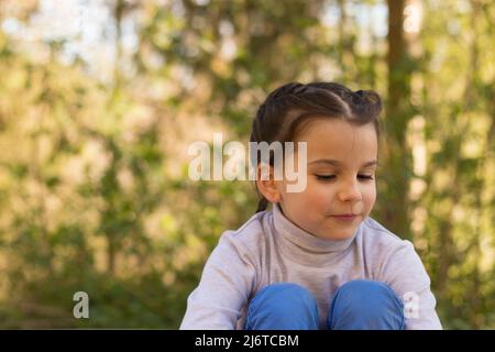 Una pequeña y hermosa colegiala sonriente sobre un fondo natural en un suéter de cuello de cisne y jeans se sienta en el suelo abrazando sus piernas Foto de stock