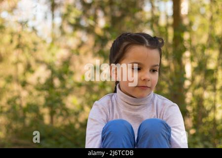Una pequeña y hermosa colegiala sonriente sobre un fondo natural en un suéter de cuello de cisne y jeans se sienta en el suelo abrazando sus piernas y mirando hacia fuera Foto de stock