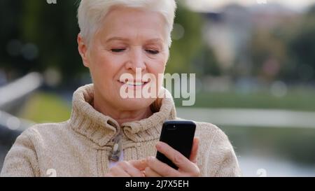 Primer plano de pelo gris Mujer madura mensajes de texto en el moderno smartphone gadget, desplazamiento, tocar la pantalla, sorprendido, abuela navegar por Internet Foto de stock