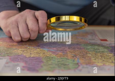 Un hombre sostiene una lupa en su mano sobre un mapa. Un hombre de mediana edad con una lupa en un marco dorado. Los nombres están escritos en ucraniano. Foto de stock
