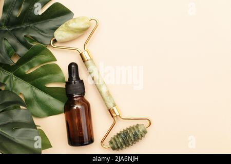 Rodillo de cuarzo de masaje para la cara de nefritis natural con aceite de suero en botella de vidrio marrón con hojas de monstera sobre fondo beige. Foto de stock