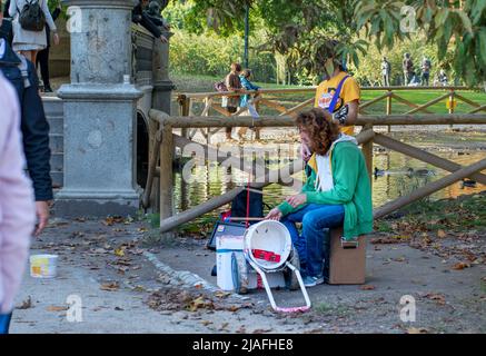 MILÁN, ITALIA - OCTUBRE 28 2016: Músicos callejeros tocan instrumentos y cantan en el parque de la ciudad. Los hombres jóvenes se sientan contra la valla de madera cerca del lago con patos adentro Foto de stock
