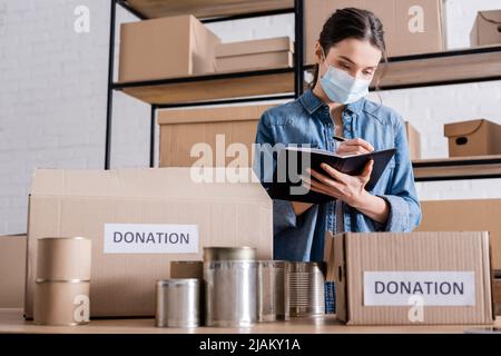 Vendedor en la máscara médica escribiendo en el cuaderno cerca de alimentos enlatados y cajas con letras de donación en la tienda web en línea Foto de stock