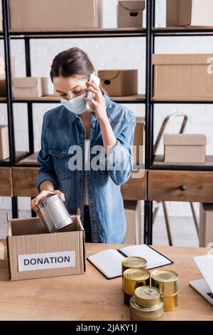 Vendedor en máscara médica hablando en el smartphone y sosteniendo comida enlatada cerca de la caja con letras de donación en la mesa Foto de stock