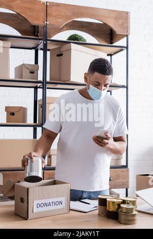Vendedor afroamericano en máscara médica sosteniendo un teléfono inteligente y alimentos enlatados cerca de la caja de donación en la mesa Foto de stock