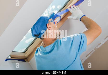 El primer plano de un trabajador masculino se centró en una aplicación cuidadosa de la cinta del pintor azul a la ventana del techo. El proceso de preparación de una sala de pintura Foto de stock