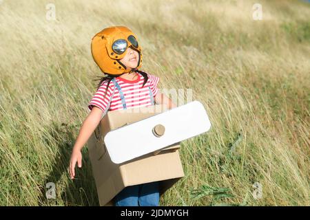 Linda chica soñadora jugando con aviones de cartón en el prado en un día soleado. Feliz niño jugando con el avión de cartón contra el cielo azul de verano ba Foto de stock