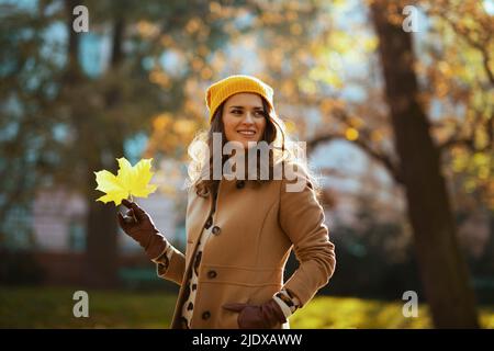 Hola noviembre. Feliz mujer moderna de mediana edad en abrigo beige y sombrero naranja con hojas amarillas de otoño al aire libre en la calle de la ciudad en otoño.