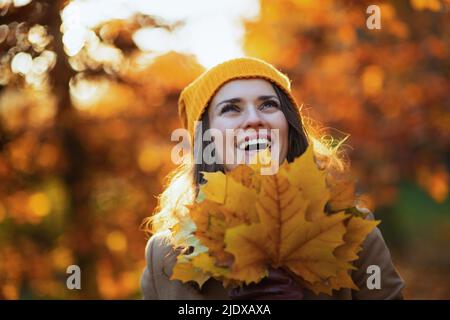 Hola otoño. Sonriente mujer de mediana edad con abrigo beige y sombrero naranja con hojas amarillas de otoño en el parque de la ciudad en otoño.
