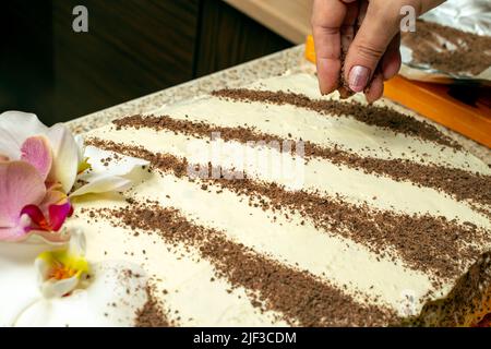 Mano de mujer rociando chocolate rallado sobre una torta blanca decorada con flores de orquídeas Foto de stock