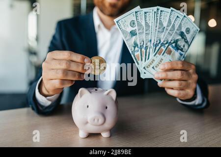 Un exitoso hombre de negocios metiendo moneda de bitcoin en el banco de cerdos y sosteniendo dinero en efectivo, sentado en la mesa del cargo Foto de stock