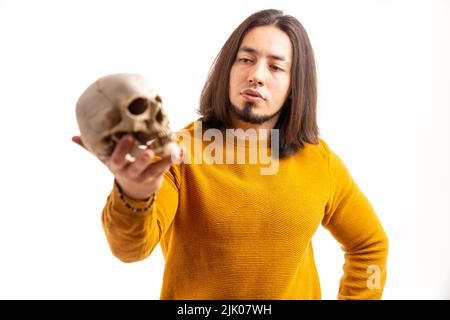 Hombre hispano barbudo con pelo largo colgando un cráneo humano blanco y mirándolo. Fotografía de alta calidad Foto de stock