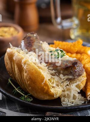 Primer plano de bratwurst a la parrilla y chucrut en un bollo con patatas fritas en un plato negro Foto de stock