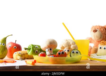 Concepto de frutas y verduras divertidas servidas en contenedores de plástico sobre una mesa de madera preparada para un niño con objetos infantiles. Vista frontal. Foto de stock