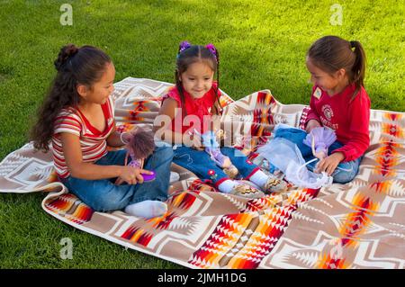 Tres chicas indígenas de edad elemental juegan juntas en una manta con muñecas Barbie. Foto de stock