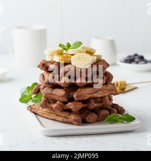 Gofres de chocolate con plátano y sirope de arce sobre mesa blanca, vista lateral. Brunch dulce, flujo de jarabe de arce Foto de stock