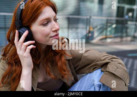 Chica joven hipster pelirroja con auriculares escuchando música en la ciudad. Foto de stock