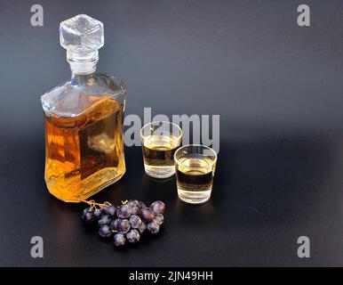 Grappa, alcohol fuerte en una botella y dos vasos sobre fondo negro, junto a un racimo de uvas oscuras. Primer plano. Foto de stock