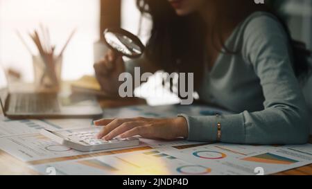 Recortada imagen de auditor femenino utilizando calculadora y examinando los datos numéricos del documento financiero mediante lupa Foto de stock
