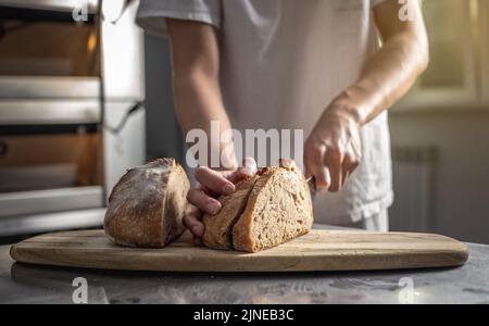 Un panadero macho corta pan recién horneado con un cuchillo para comprobar la calidad. Producción de productos de panadería como pequeña empresa. Foto de stock