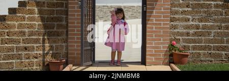 Imagen de una adorable niña en un delantal rosa y una mochila que saluda a sus padres en su primer día de escuela. Banner horizontal Foto de stock