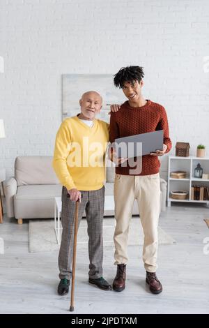 Jubilado sonriente con bastón mirando la cámara cerca de un nieto afroamericano con un portátil en casa Foto de stock