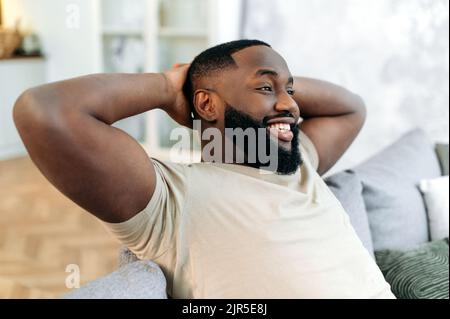 Relajado, satisfecho y tranquilo hombre afroamericano con ropa informal, descansando en un cómodo sofá en un acogedor salón, tirando las manos detrás de su cabeza, soñando con vacaciones, descansando del trabajo, sonriendo Foto de stock