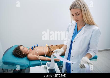 Pediatra buscando impresión de ECG de niño paciente después de un procedimiento de electrocardiograma del corazón en la clínica médica Foto de stock