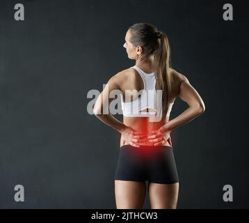 Sentirlo en su espalda. Fotografía de estudio de una mujer joven y deportiva con dolor en la parte baja de su espalda contra un fondo oscuro. Foto de stock