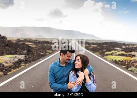 Hombres y mujeres jóvenes viajeros abrazaron y caminaron a lo largo de carreteras asfaltadas en terreno montañoso durante sus vacaciones en Lanzarote Foto de stock
