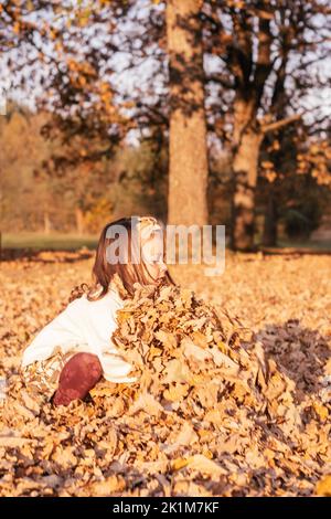 Niña de 3-4 años de edad cuclillas en otoño al aire libre en el parque en los rayos de puesta de sol y recoger grandes armaduras de hojas de roble. Niño de pelo oscuro adentro Foto de stock