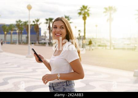 Millennial niña caminando sosteniendo un smartphone al atardecer en el paseo marítimo de la ciudad Foto de stock