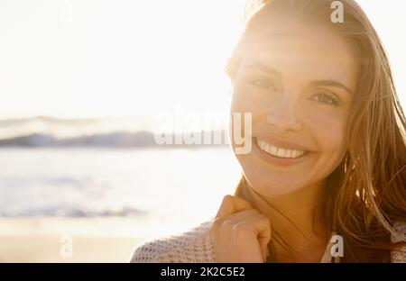 Una sonrisa impresionante al atardecer. Una mujer joven y bonita sonriendo con confianza mientras el sol se pone en la playa en el fondo. Foto de stock