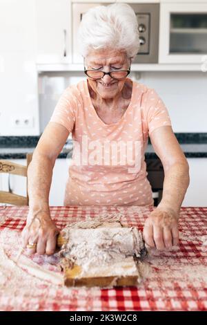 Alegre anciana con pelo gris en gafas rodando masa sobre tabla de cortar en la mesa de casa Foto de stock