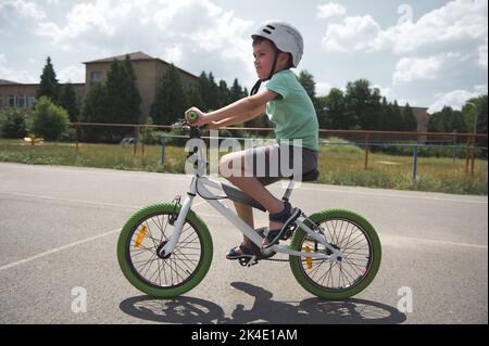 Estudiante de escuela caucásico, deportivo y activo, con casco deportivo en bicicleta por la carretera asfaltada en un entorno urbano Foto de stock