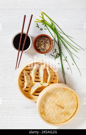 gyoza, wonton envoltorios rellenos de cerdo y col en vaporera de bambú sobre mesa de madera blanca con salsa de soja y cebolletas, vista vertical desde arriba Foto de stock