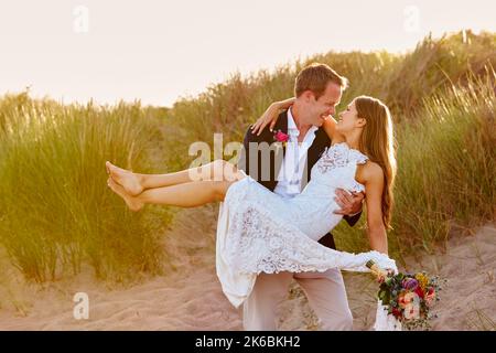 Romántica pareja de casados celebrando boda en la playa con novio llevando a novia en las dunas