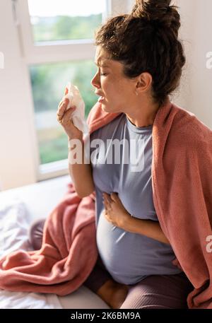 Mujer embarazada cubierta con una manta sentada en la cama y estornudando en un pañuelo. Concepto de frío o virus en período de maternidad Foto de stock