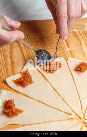 La mano pone una cuchara de mermelada de manzana en los triángulos de la masa para la preparación de croissants franceses. Vista superior. Concepto de horneado casero Foto de stock